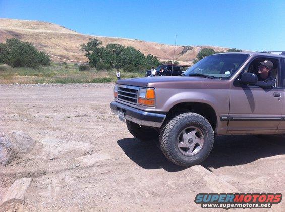 Brown Ford Explorer on a dirt road, driver visible. Another vehicle and people in background