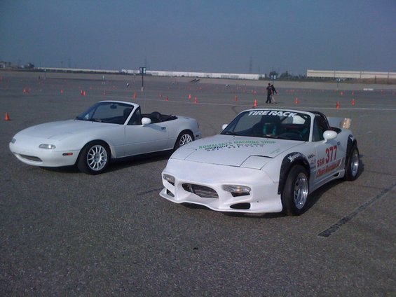 Two white Mazda Miatas parked on asphalt with orange cones in the background. One has racing decals