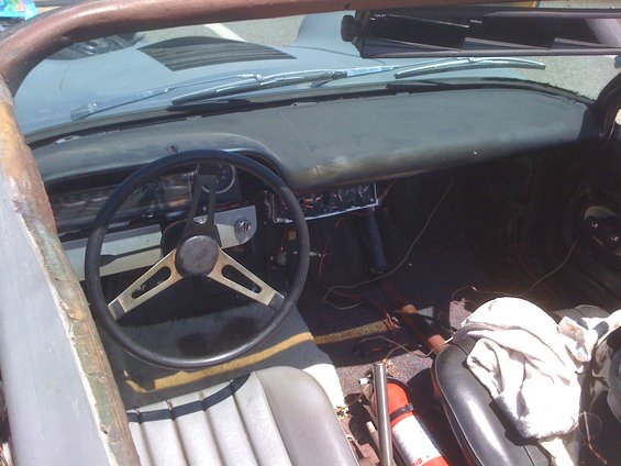 Interior of a car with a vintage steering wheel, dashboard, and seats. Wires are visible, and a fire extinguisher is on the floor