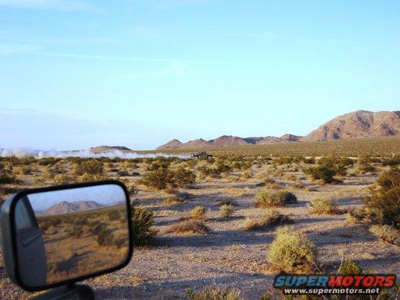 Truck kicking up dust in a desert landscape, seen from a side mirror