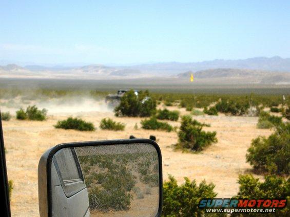 Two off-road vehicles driving through a dusty desert landscape with sparse vegetation and distant mountains