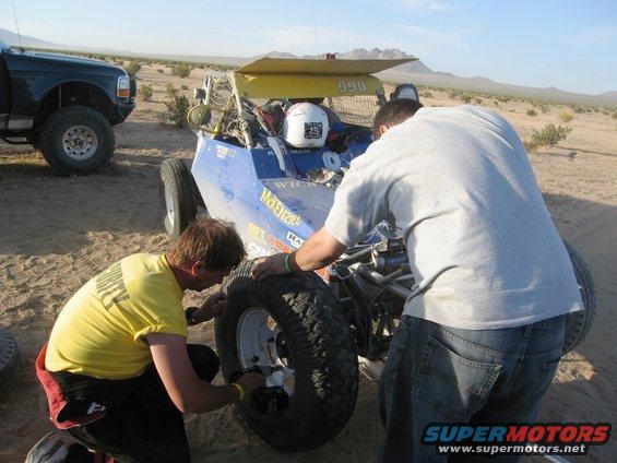 Two men working on a blue off-road buggy with yellow wing in desert; dark pickup truck in background