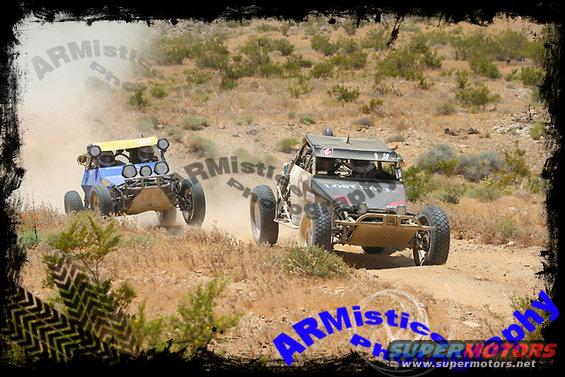 Two off-road buggies kick up dust while racing on a dirt track in a desert landscape