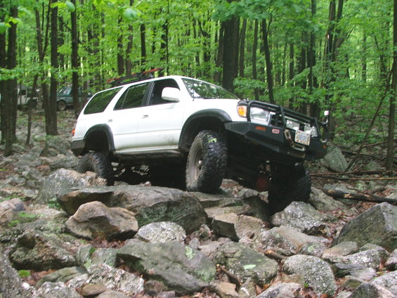 White Toyota 4Runner with aftermarket bumper and large tires navigating rocks. Another vehicle is visible in the background