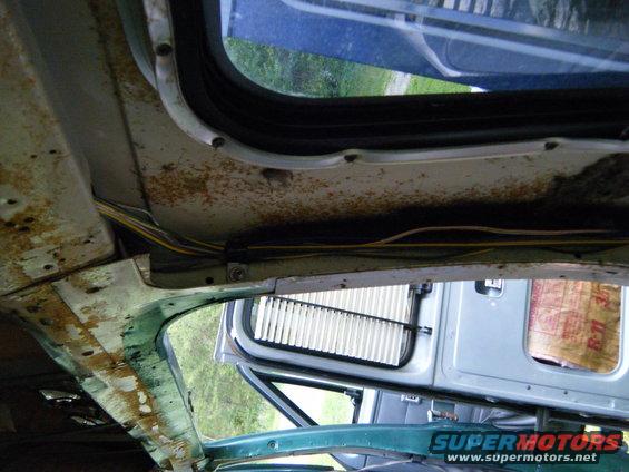 View of the rusty interior ceiling and window frame of an old van. Wires are visible. Another vehicle's door is seen through the window