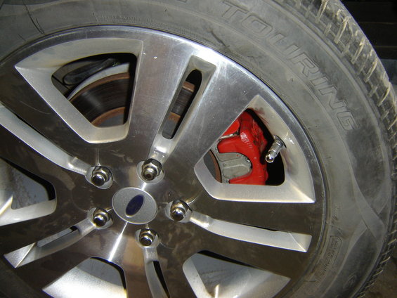 Close-up of a car wheel with a red brake caliper visible through the spokes. Tire has 'TOURING' text