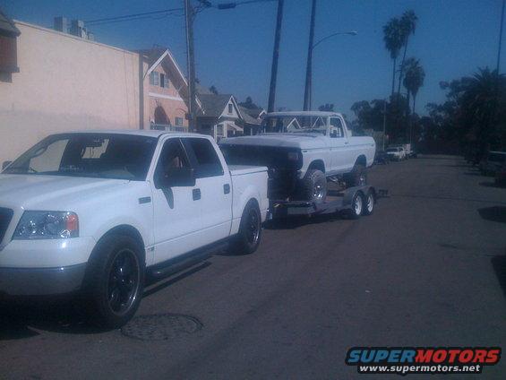 White pickup truck towing a white 1970s pickup truck on a trailer, side view