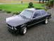1982 BMW 3 Series - A black BMW sedan with gold wheels is parked on a gravel driveway in front of a lawn and trees.