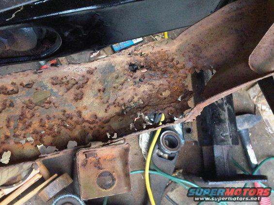 Heavily rusted frame rail with visible holes and peeling paint. Tools and wiring are present