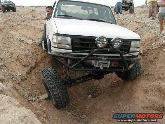 White Ford Bronco with off-road modifications navigating a deep rut, front-end visible