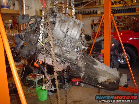 Engine and transmission assembly suspended by chains in a garage. A red vehicle is partially visible in the background