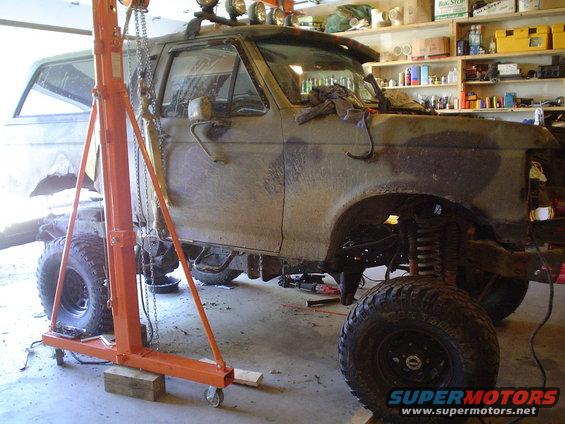 Mud-splattered 1990 Ford Bronco with an engine hoist, lifted suspension, and off-road tires in a garage