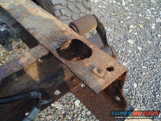 Rusted frame section with a large hole, showing suspension components and wiring