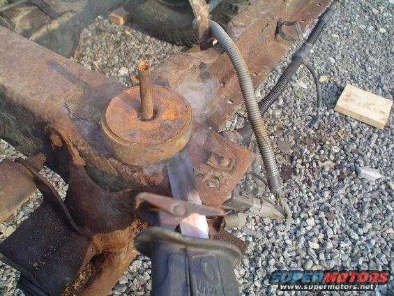 Cutting a rusty metal frame component with a reciprocating saw. Gravel and some hoses are visible