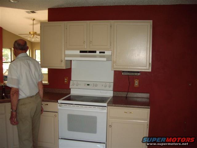 Man in a white shirt and tan pants stands in a kitchen, facing away from the camera. A white stove and cabinets are visible