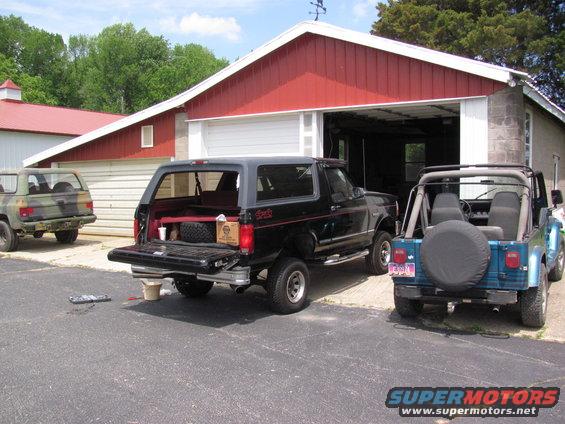 1995 Ford Bronco with tailgate open, parked next to two Jeeps by a red garage