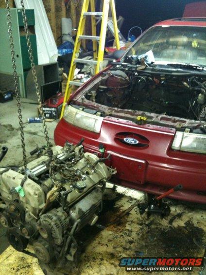 red car with hood open, engine removed and hanging from a chain hoist, ladder in background