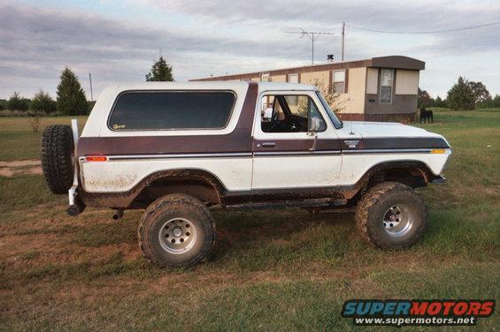 White and brown Ford Bronco parked on grass with a spare tire on the back. A trailer home is in the background