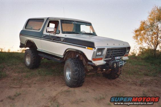 White and dark green 1978 Ford Bronco with large tires and a winch, parked on a dirt incline
