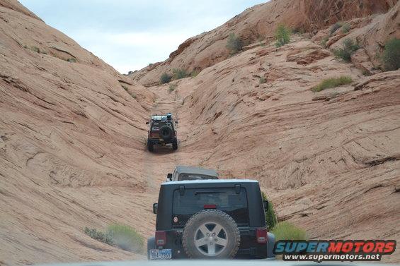 Black Jeep Wrangler rear view, spare tire mounted, driving on dirt road between sandstone cliffs