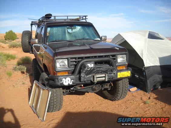 Brown Ford Bronco II with a roof rack and spare tire on the back, parked next to a tent in a desert landscape