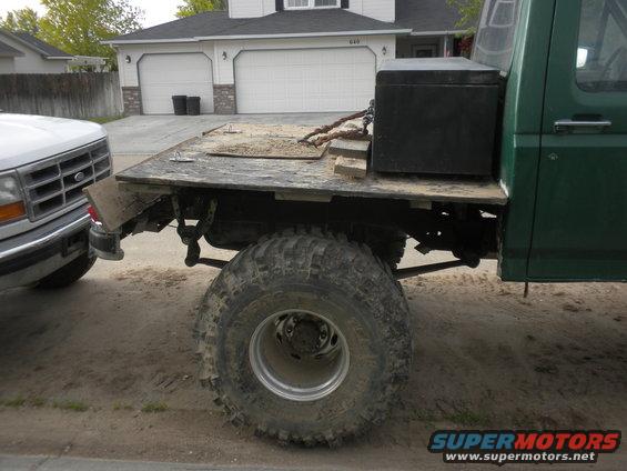 Green flatbed truck with large tires, next to a white pickup truck. Toolbox on the flatbed