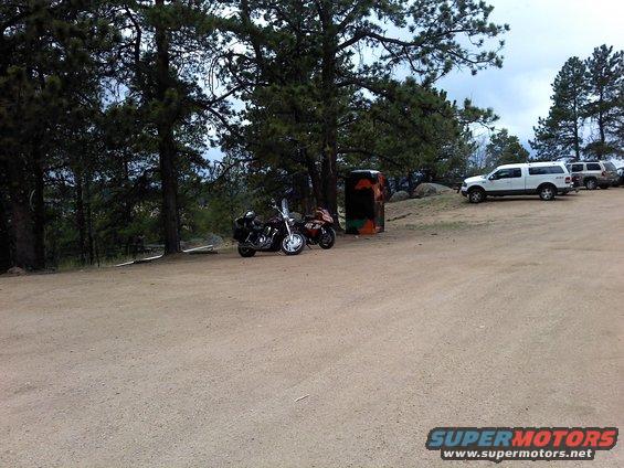 Two motorcycles parked near trees, with SUVs in the background on a dirt lot