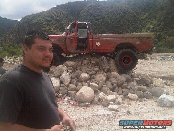 Man in foreground, red pickup truck with open door perched on a pile of rocks