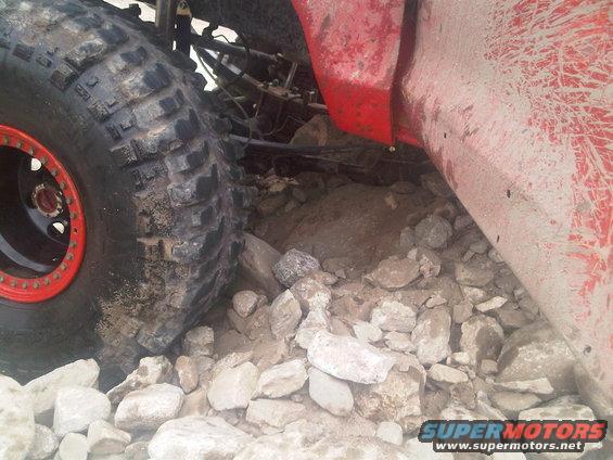Close-up of a large off-road tire with a red beadlock ring, resting on rocks. Red body panel visible