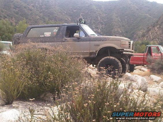 Muddy 1992 Ford Bronco with off-road lights and spare tire on rear, parked on rocky terrain next to a red pickup truck
