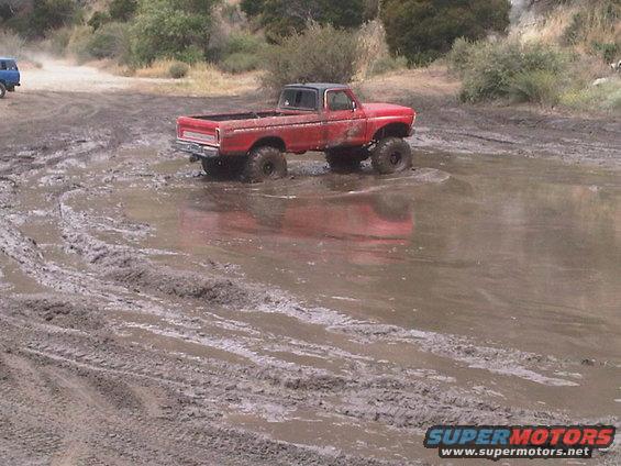 red pickup truck with big tires stuck in mud puddle, another blue vehicle in background