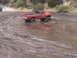 1986 Ford Bronco - A red pickup truck navigates a muddy off-road course, with another vehicle visible in the distance.