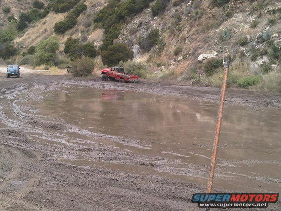 Red pickup truck stuck in mud pit; another vehicle visible in background