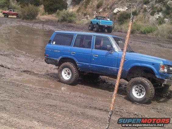 Blue Toyota Land Cruiser FJ60 with lifted suspension driving through mud, red pickup and blue Jeep in background