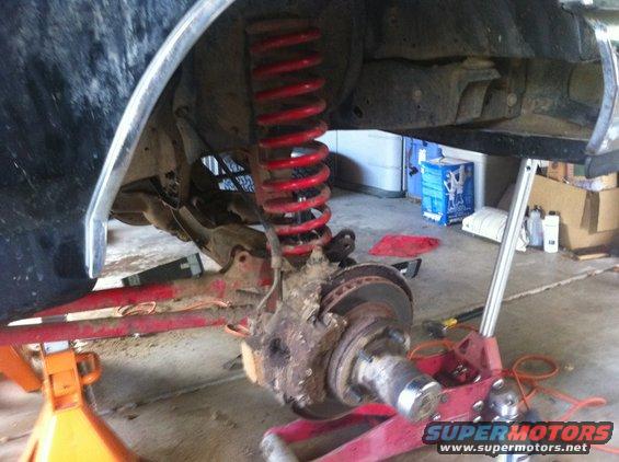 Vehicle's front suspension and brake assembly on jack stands, showing red coil spring and rusty components