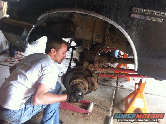 Man working on front suspension of a lifted Ford Bronco, which has a 'Bronco' badge on the fender