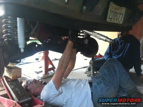 Undercarriage of a lifted 1990 Ford Bronco with a mechanic working on the rear axle and another person watching