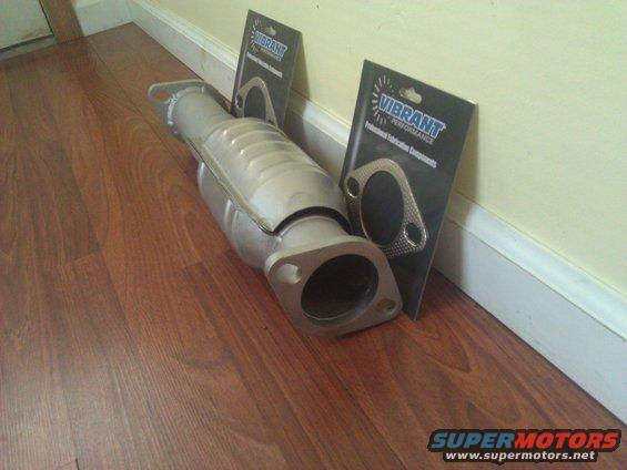 Catalytic converter and two Vibrant Performance gaskets leaning against a wall on a wood floor