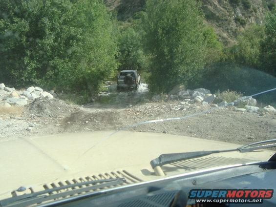 View from inside a vehicle looking at another SUV driving through a shallow stream in a wooded area