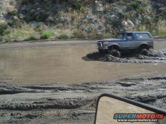 Gray and blue Ford Bronco driving through mud, lifted suspension, off-road tires