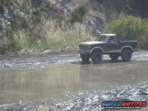 Tan and dark blue 1980s Ford Bronco driving through mud