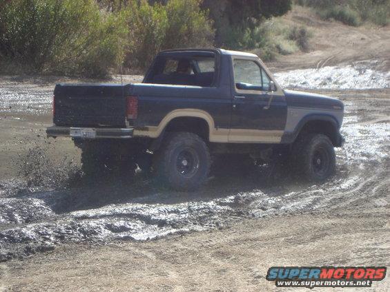 Ford Bronco driving through mud, kicking up spray. Two-tone paint, lifted suspension