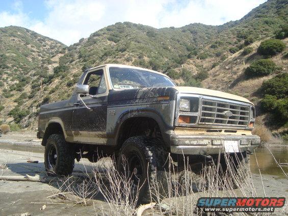 Ford Bronco parked in dry riverbed with muddy tires and body, hills in background