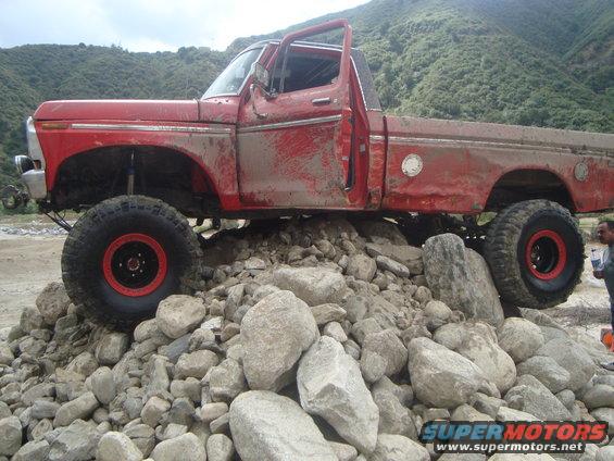 Red pickup truck with large tires is driving over a pile of rocks. The truck is muddy and has its doors open