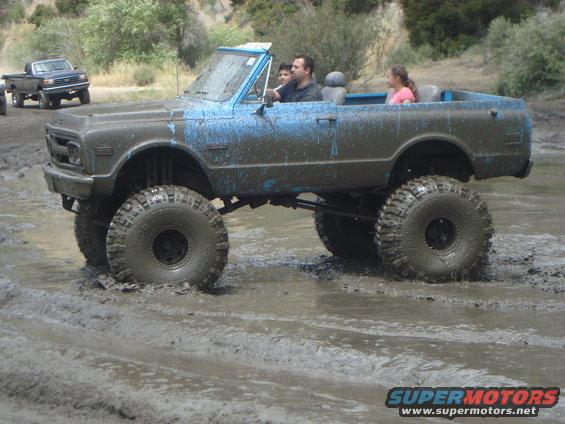 Mud-covered lifted Chevrolet K5 Blazer with large tires driving through mud, blue paint visible under the mud