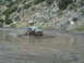 1986 Ford Bronco - A dark SUV navigates a deep mud puddle on a rough, unpaved road.