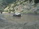 1986 Ford Bronco - A tan pickup truck navigates through a muddy puddle on a dirt road, with another vehicle visible in the distance.