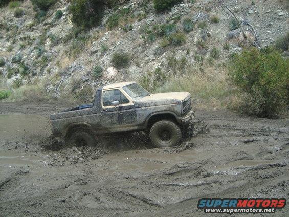 Muddy 1980s Ford Bronco driving through a deep mud puddle on a dirt road