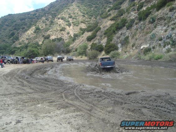 Blue pickup truck splashing through a muddy puddle with other vehicles and people in the background