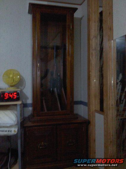 wooden gun cabinet with glass doors, digital clock showing 9:45, and a yellow fan